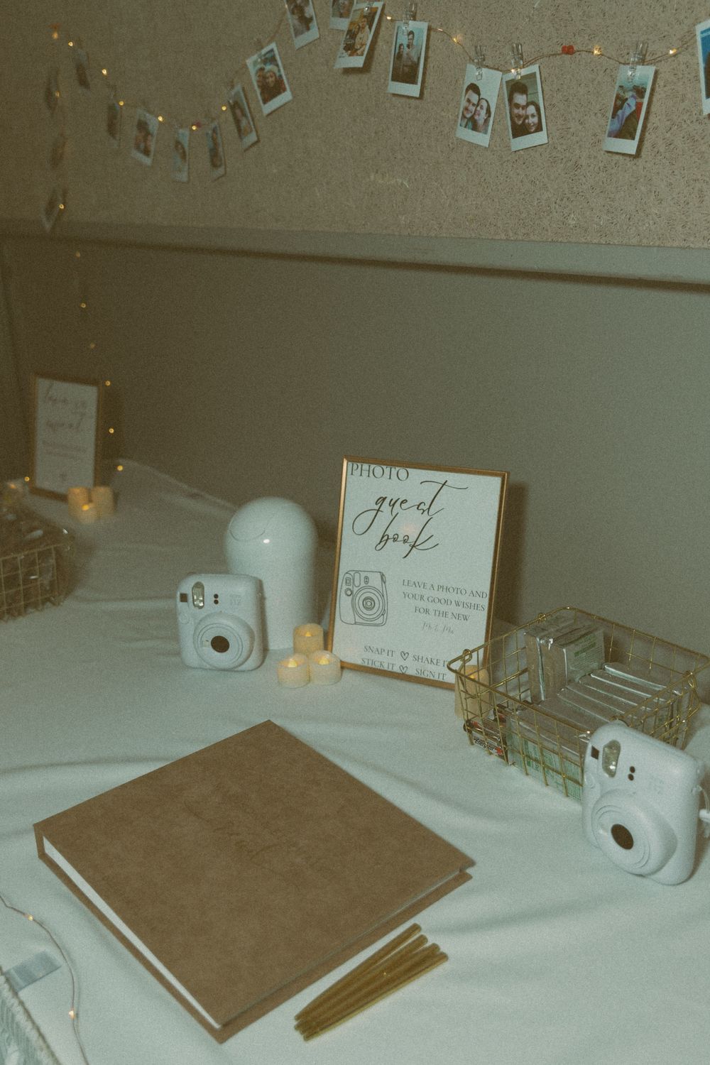 Vintage-style wedding guest book table setup with polaroid cameras and string lights on white tablecloth.