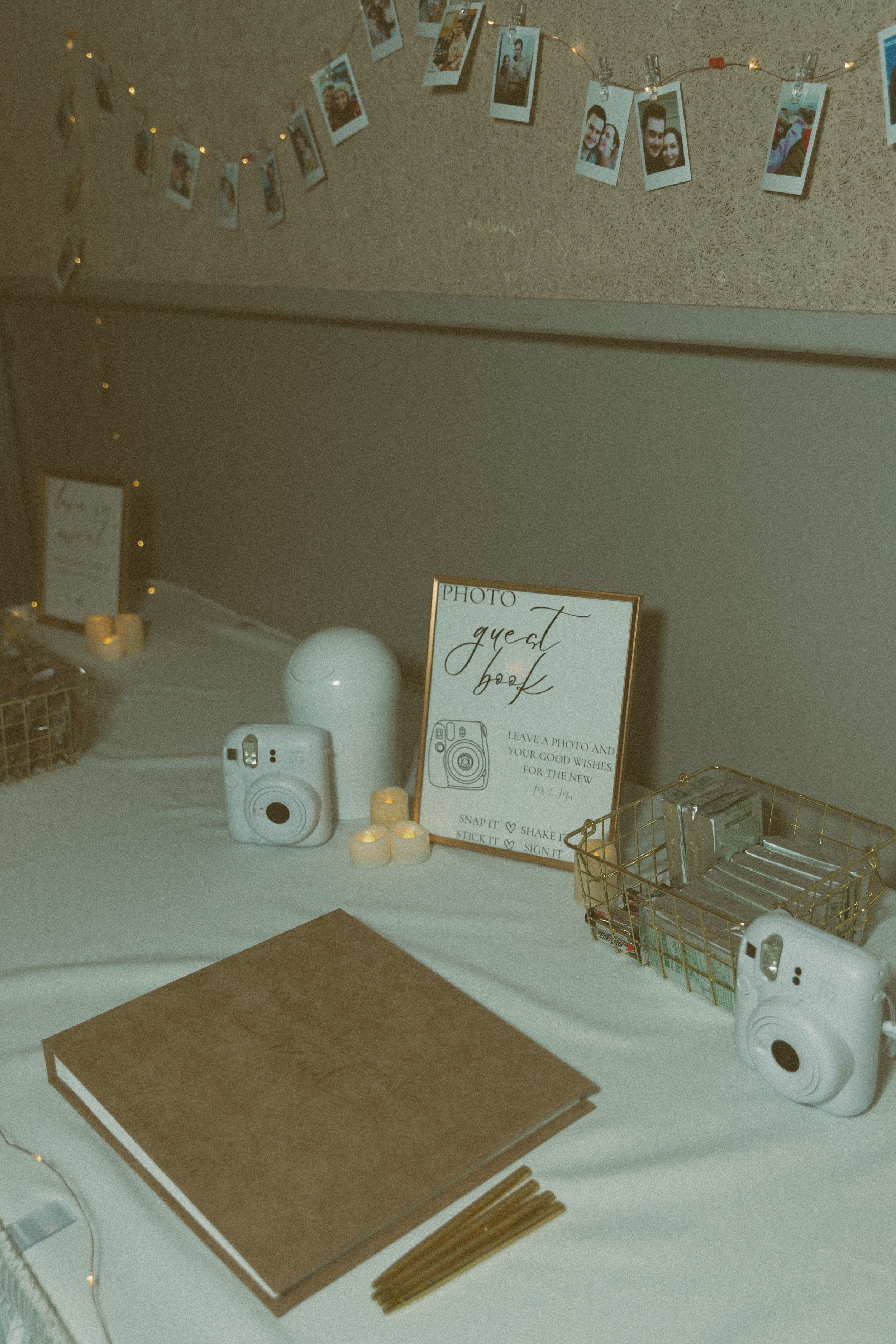 Vintage-style wedding guest book table setup with polaroid cameras and string lights on white tablecloth.