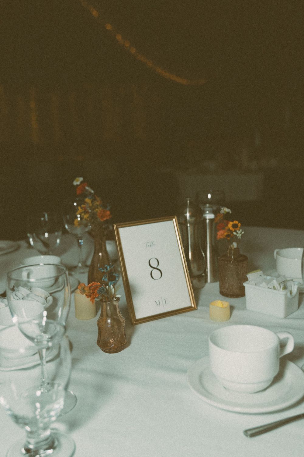 A vintage-style table setting with a framed number 8, white teacups, water glasses, and small amber flowers in vases.