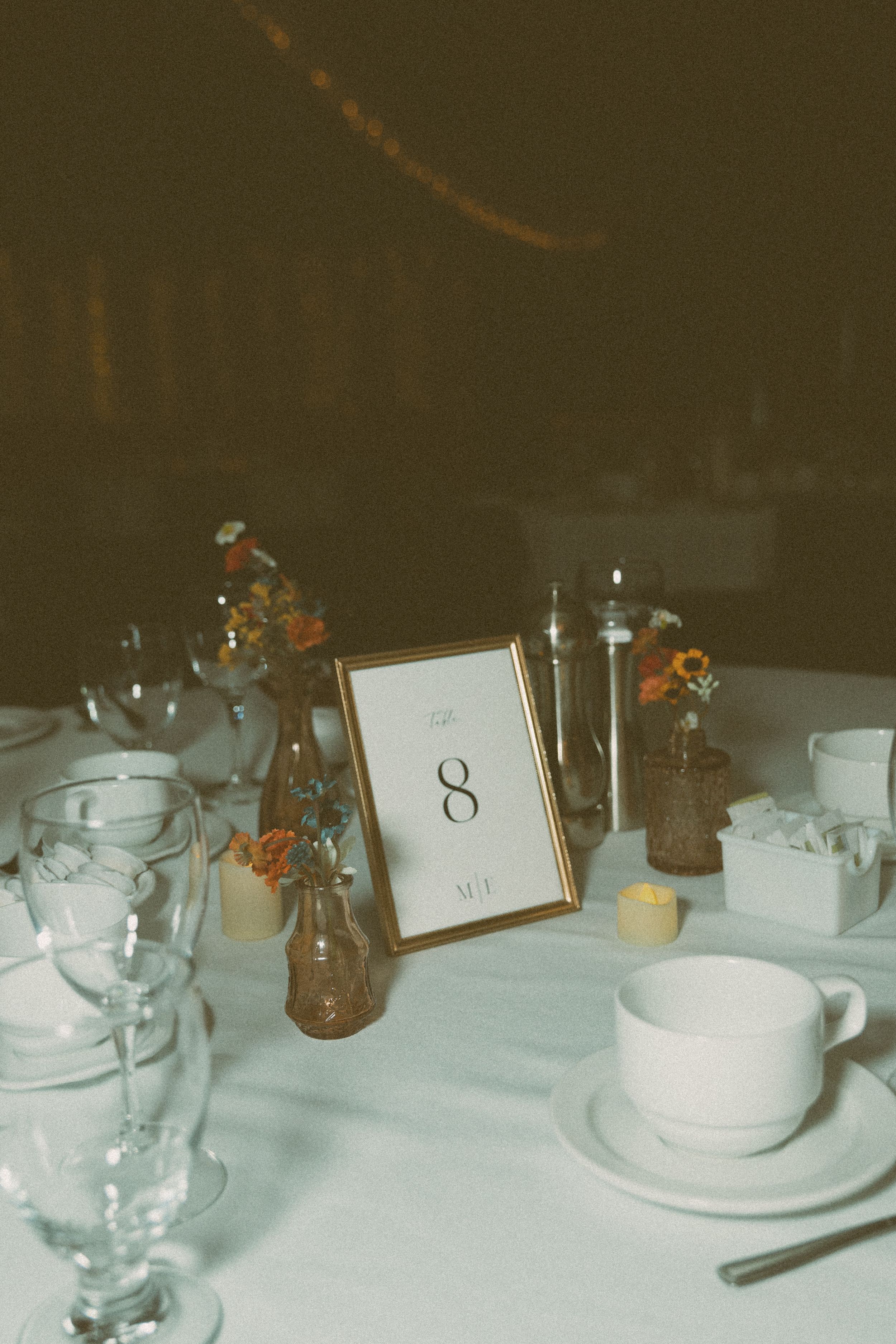 A vintage-style table setting with a framed number 8, white teacups, water glasses, and small amber flowers in vases.