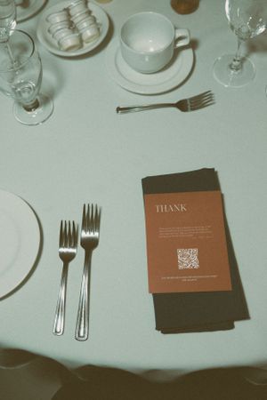 Elegant place setting at wedding reception with white dishes, silverware and thank you card on tablecloth.