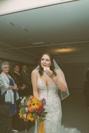 A bride in a lace wedding dress holds a vibrant sunflower bouquet while others look on in the background.