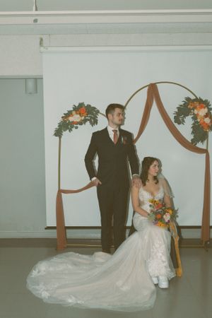 A wedding couple poses under an arch decorated with orange flowers and brown draping fabric at their indoor ceremony.