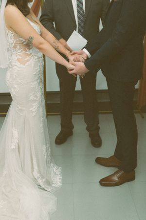 Close-up view of hands joined during wedding ceremony with bride in lace gown and groom in suit with brown dress shoes.