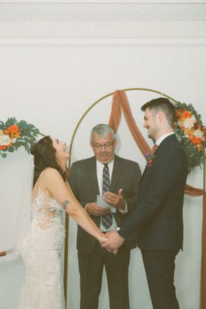 A couple sharing a lighthearted moment during their wedding ceremony under an arch decorated with orange flowers and ribbon.