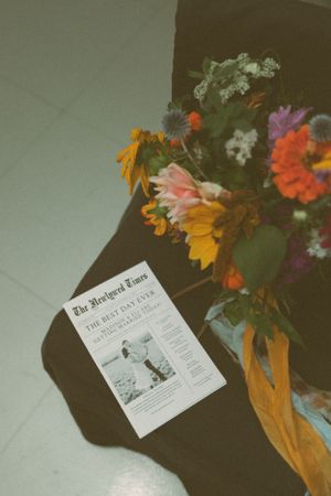 A newspaper and colorful floral arrangement with sunflowers and orange ribbons on a dark surface.