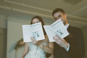 A couple holds up wedding event programs while posing together in a dreamy film-style photo.