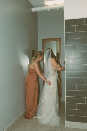 A bridesmaid in a rust-colored dress helps adjust the train of a white lace wedding gown in a modern hallway.