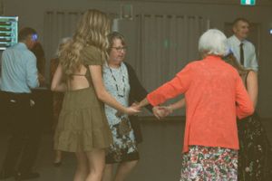 People dance together at a social gathering with a woman in an orange cardigan and floral skirt visible.