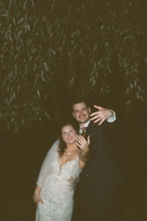 A couple poses and waves under twinkling lights at an evening outdoor celebration.