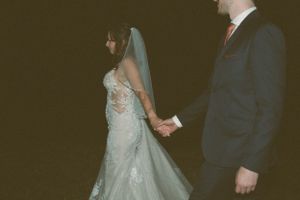 A wedding couple in formal attire stands together during their ceremony against a dark backdrop.