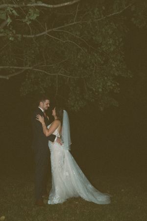 A wedding couple embraces under tree branches at night in a romantic outdoor setting.