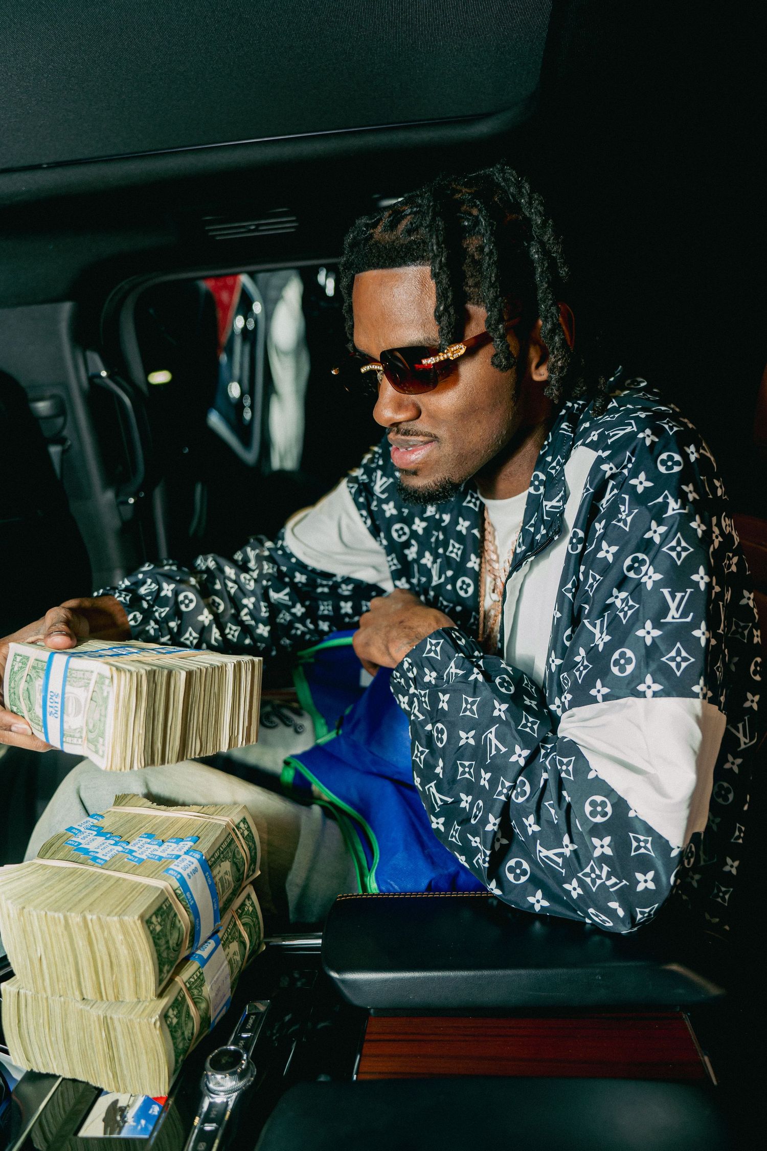 Person wearing Louis Vuitton jacket and sunglasses sits in car counting stacks of cash money.