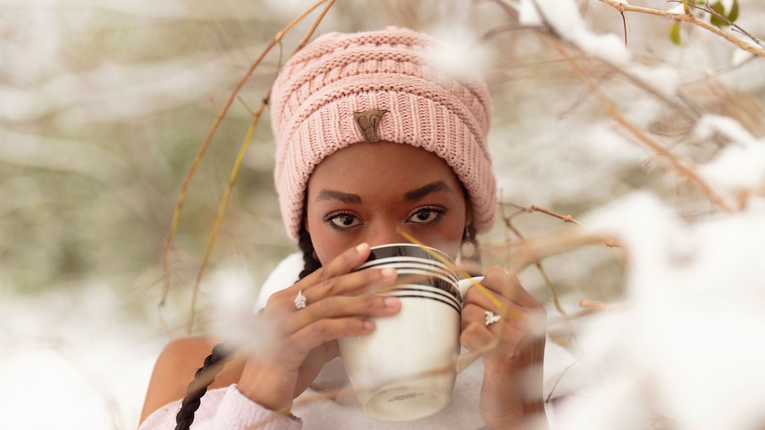 Someone wearing a pink knit beanie drinks from a white mug outdoors on a bright winter day.