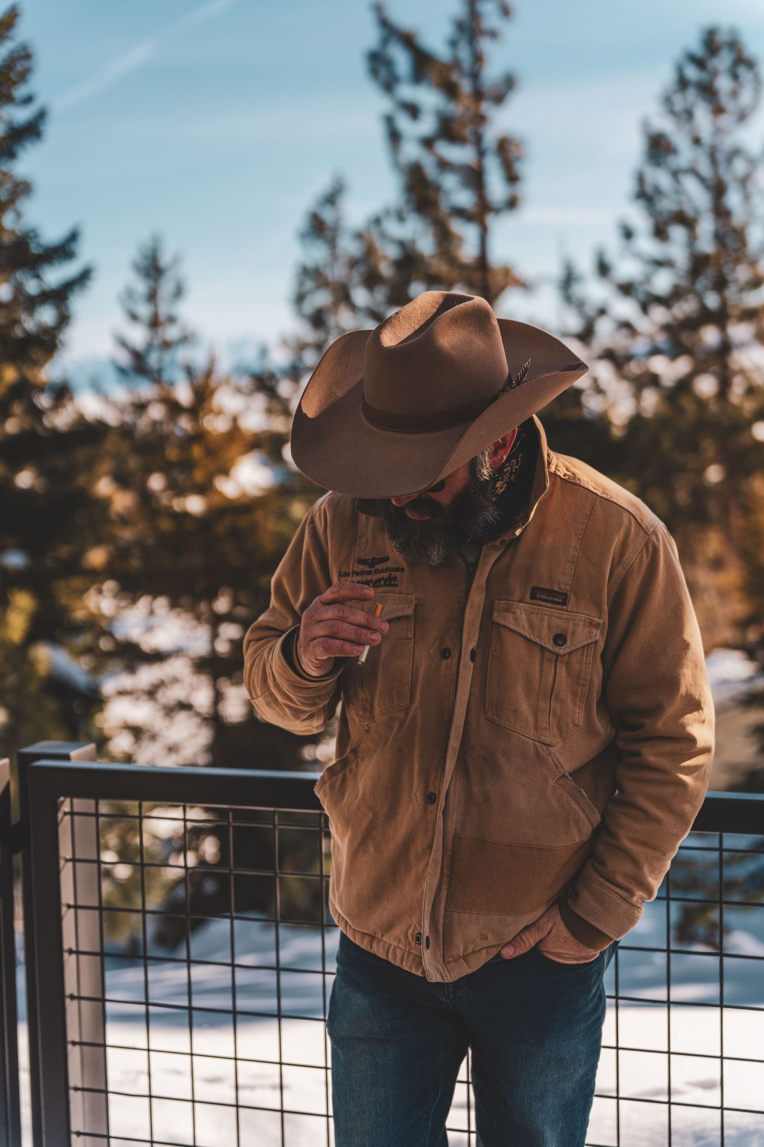 A rugged figure in a brown cowboy hat and canvas jacket stands against a snowy winter forest backdrop at sunset.