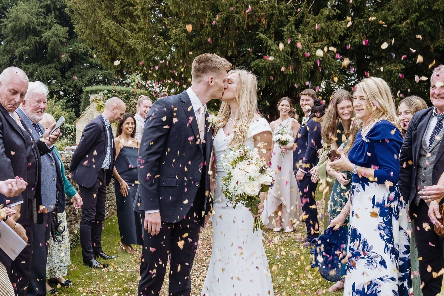 Wedding guests throw confetti over newlyweds during outdoor ceremony celebration with trees in background.