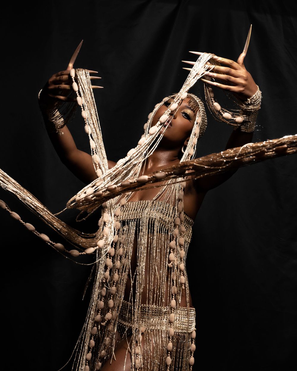 Avant-garde fashion photo featuring model in metallic fringe dress with dramatic arm poses.