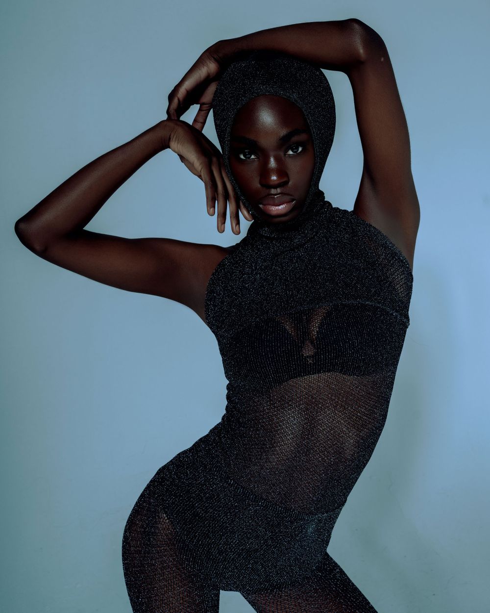 Fashion model poses in black mesh bodysuit against blue backdrop in dramatic studio portrait.