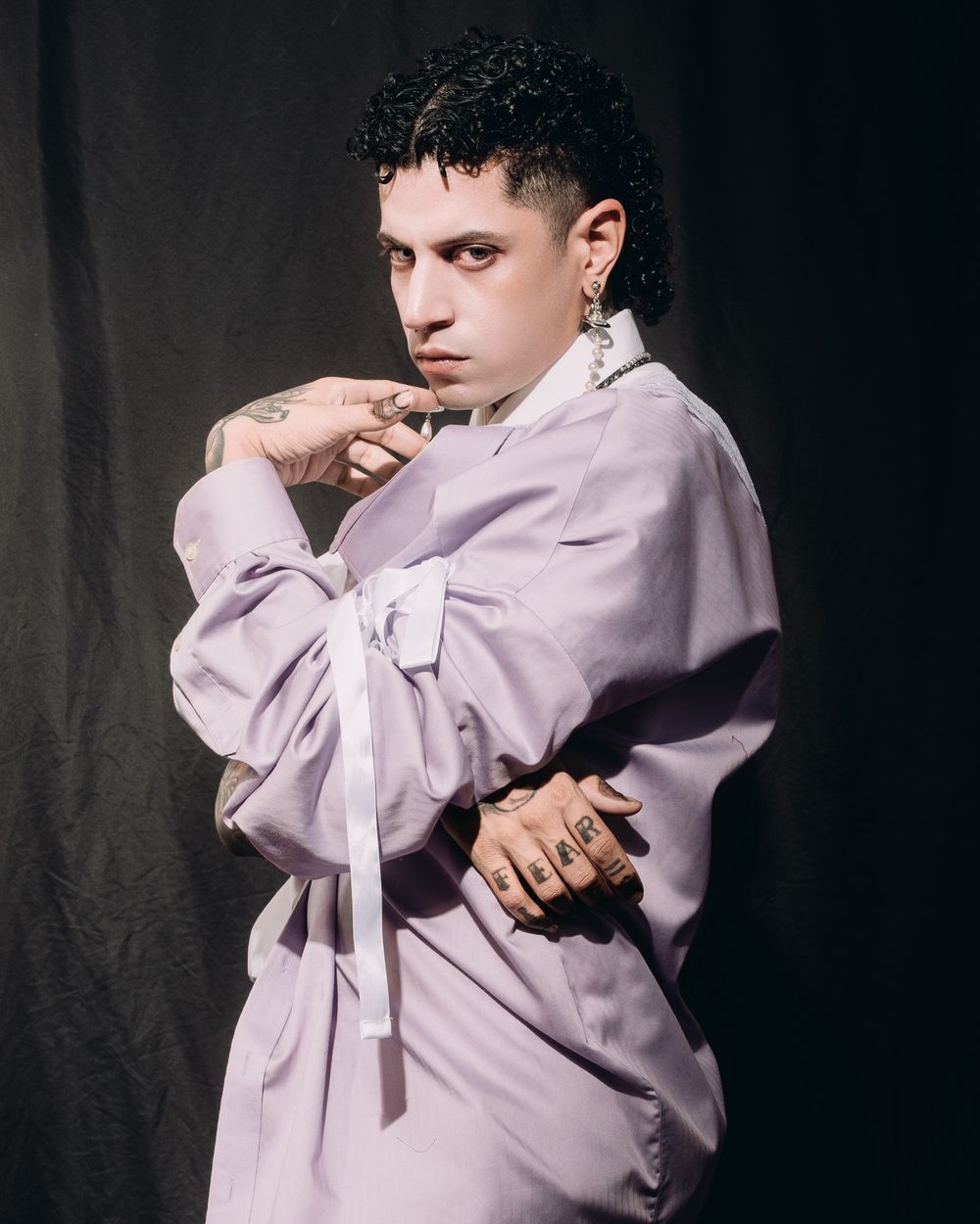 Portrait against dark backdrop featuring figure in oversized lavender button-up shirt with dramatic styling and jewelry.