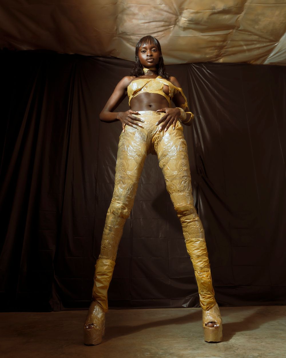 Fashion model in metallic gold outfit posing dramatically against dark backdrop.