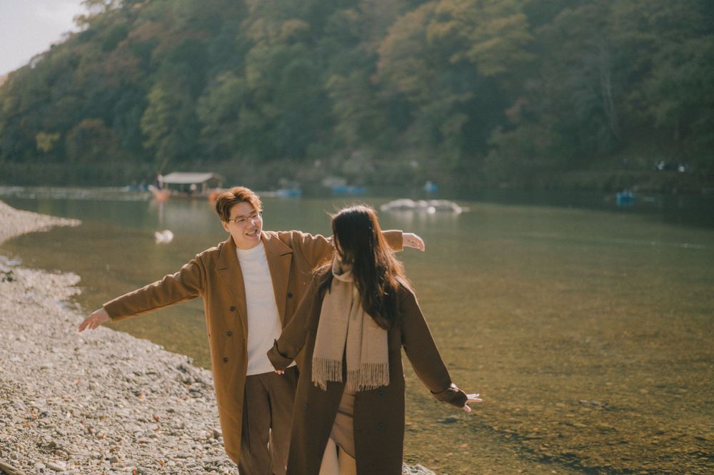 Couple walking hand in hand along a scenic riverside shore with lush green hills behind.