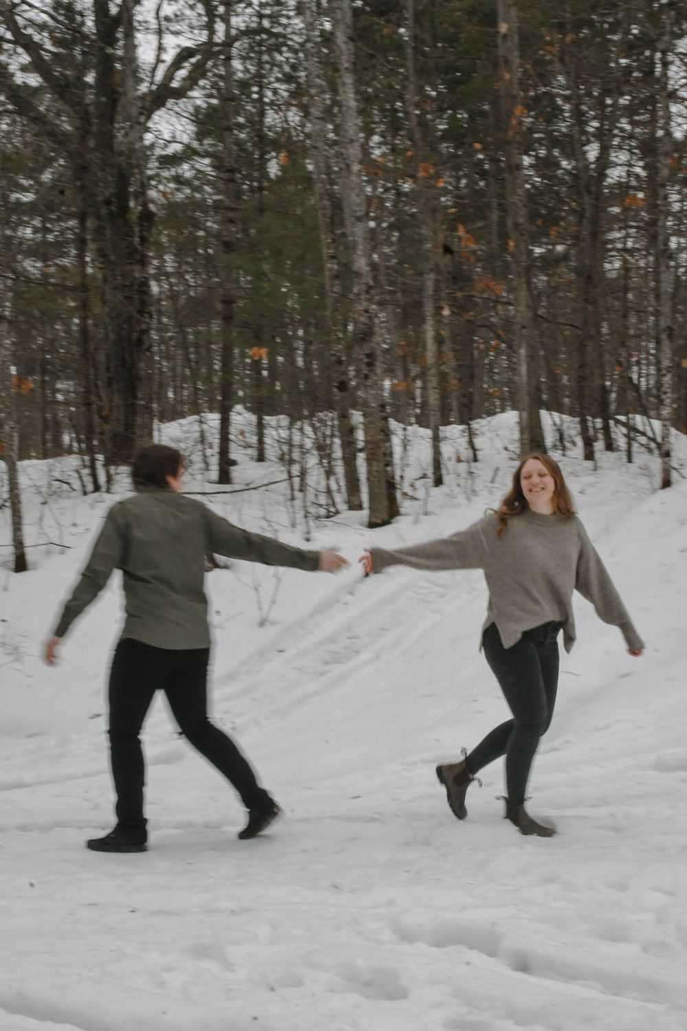 A couple dances together on a snowy trail through a winter forest in casual sweaters.