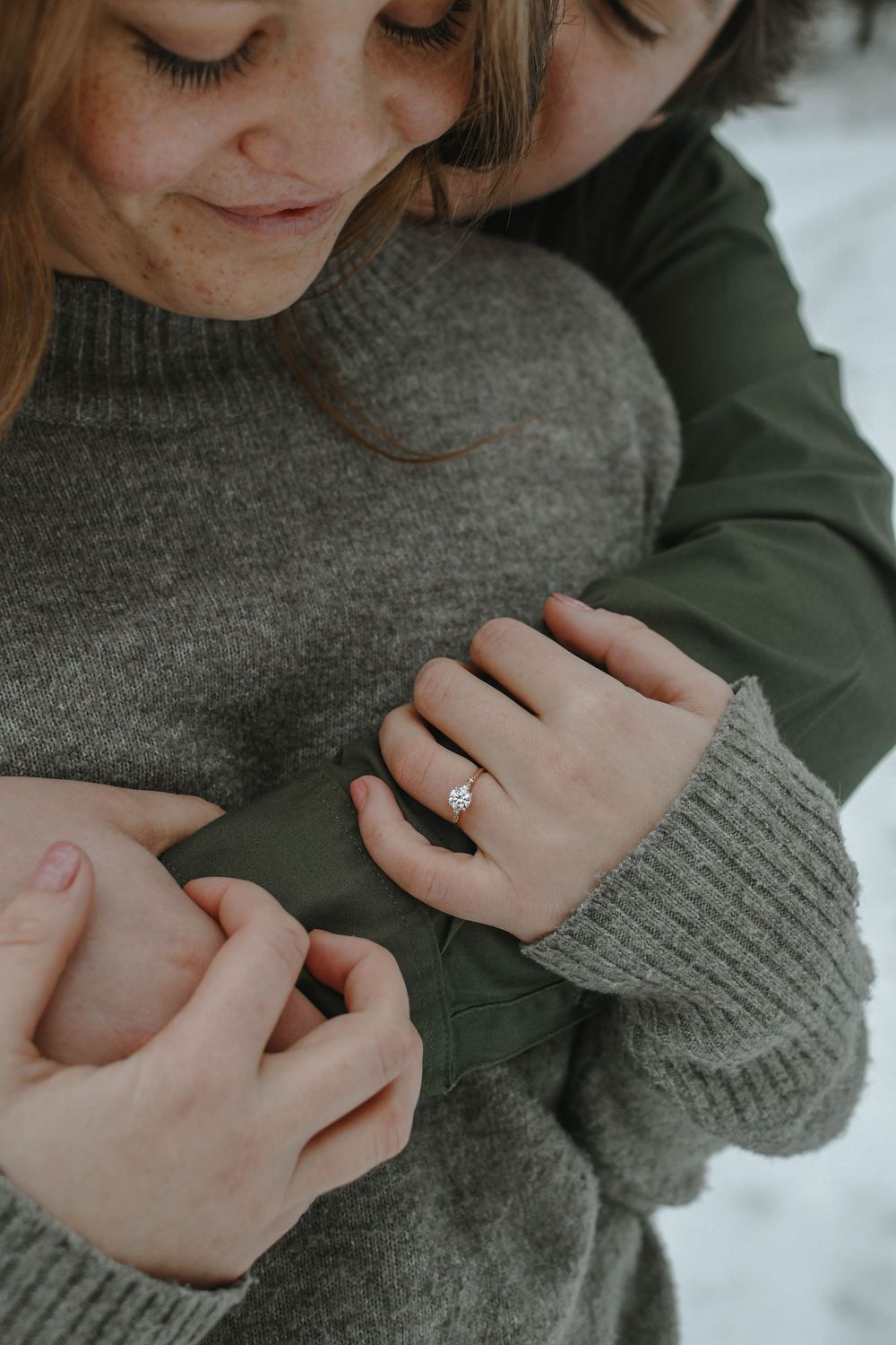 Close-up of two people embracing, wearing gray sweater and green jacket, with engagement ring visible on hand.