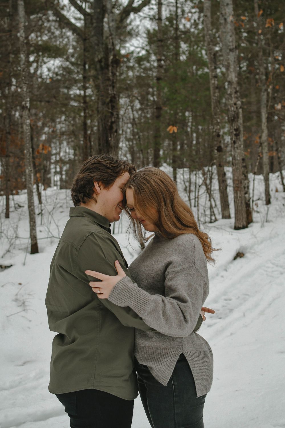 A romantic winter scene in a snowy forest shows a couple embracing while wearing cozy sweaters.