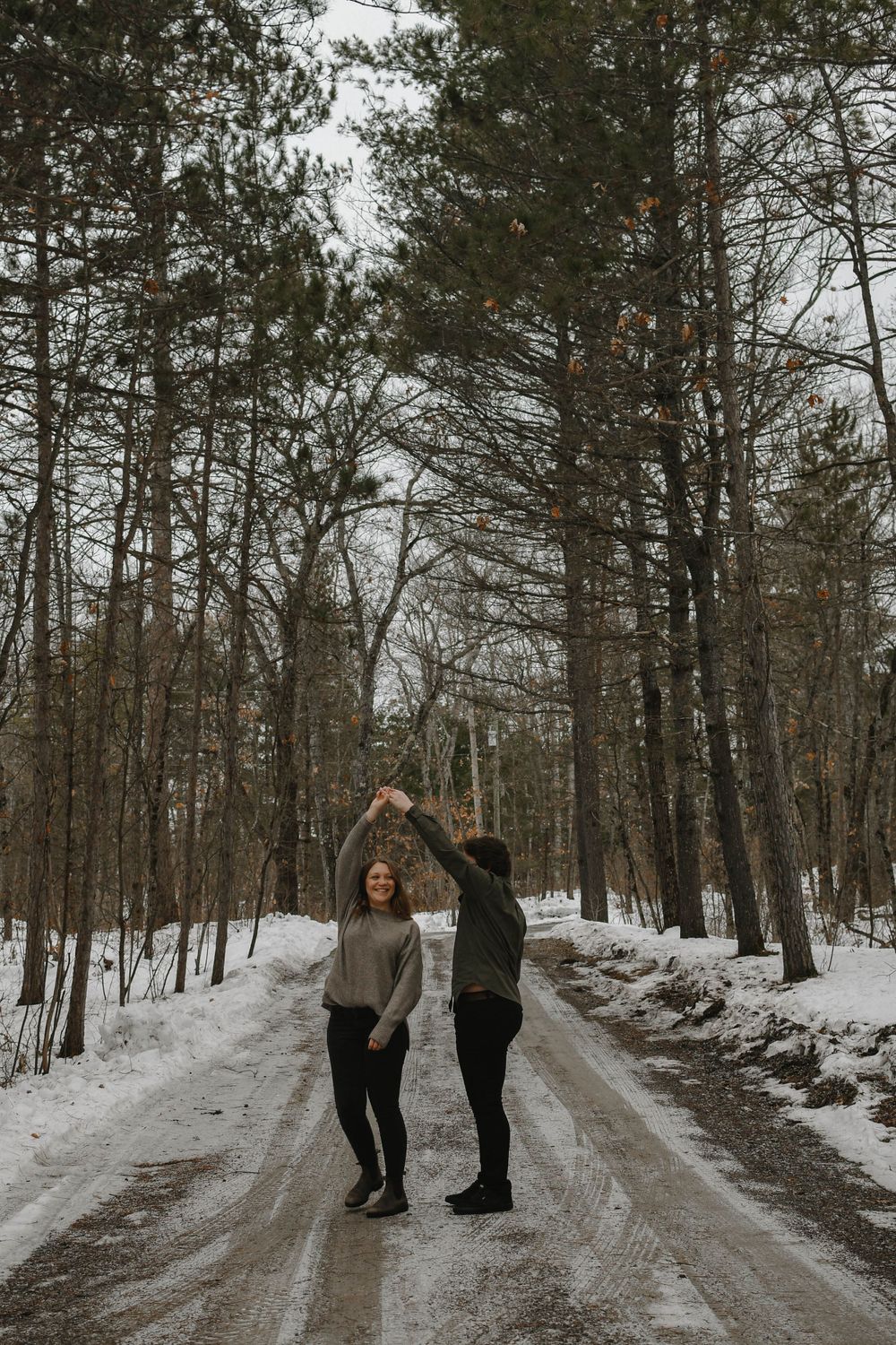 Two people dancing playfully on a snowy forest path surrounded by tall pine trees on a winter day.