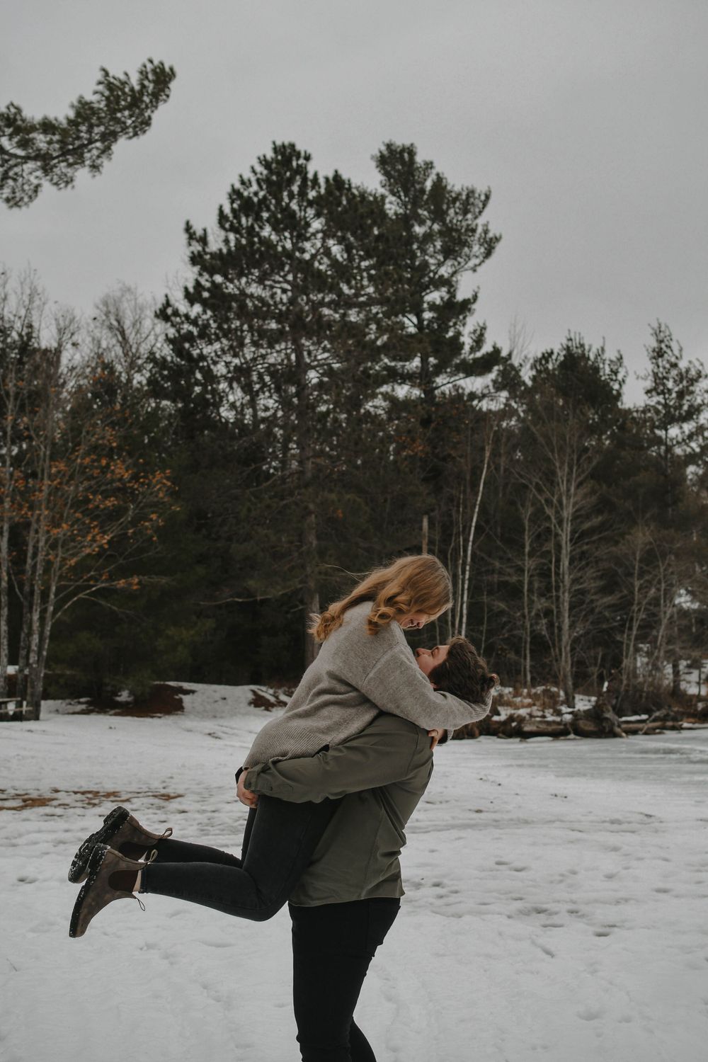 A couple embraces while standing in the snow with tall pine trees in the background on a winter day.