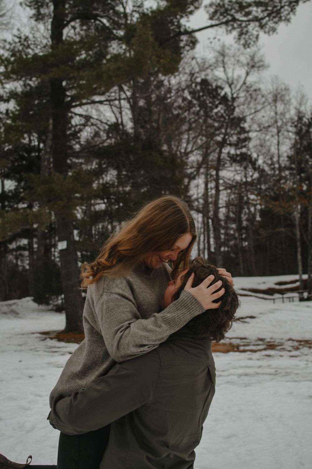 Two people share a romantic winter moment embracing in a snowy forest setting with tall pine trees in the background.