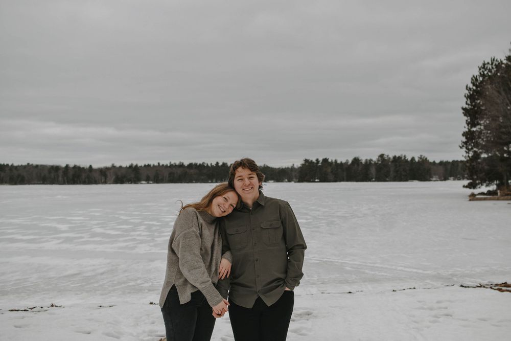 Two people embrace while standing on a snow-covered lake shore with evergreen trees and overcast sky in the background.