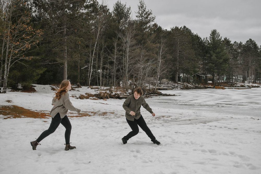 Two people run playfully through a snowy winter landscape with pine trees and melting patches of snow in the background.
