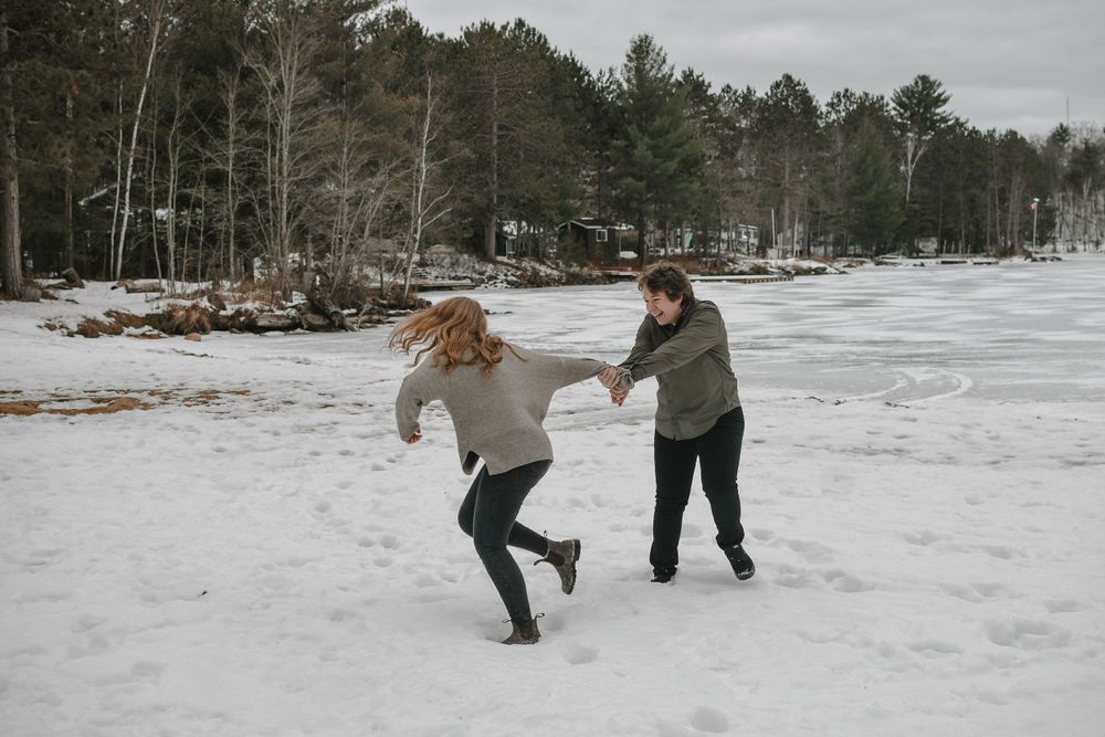 Two people playfully dance together on a snowy winter day with pine trees and frozen lake in background.