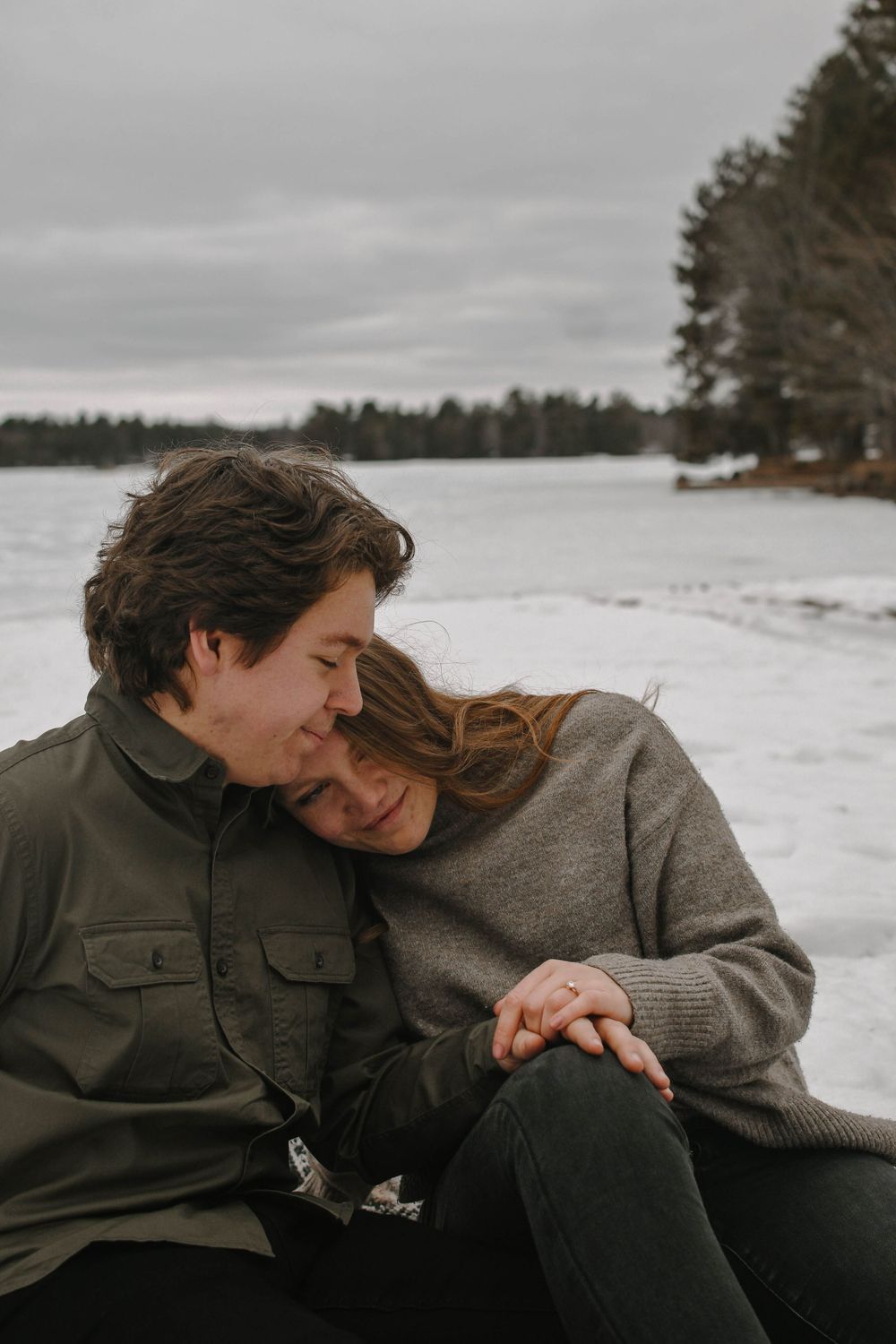 A couple shares an intimate moment while sitting together on a snowy winter day by a lake.