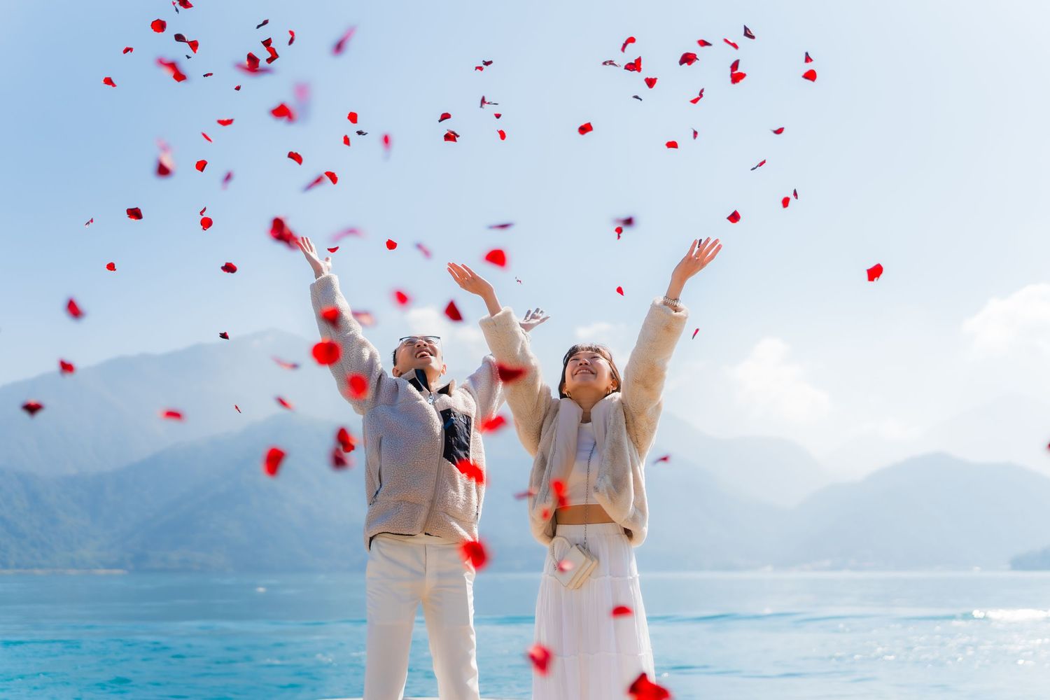 A couple celebrates engagement and proposal by throwing red rose petals in the air near a bright blue lake, Sun Moon Lake in Taichung Taiwan