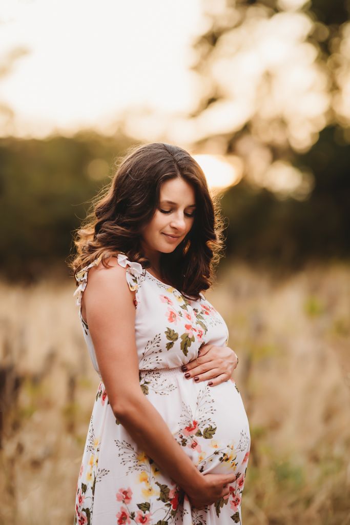 Expectant mother in a floral dress poses in a sunlit field during golden hour.