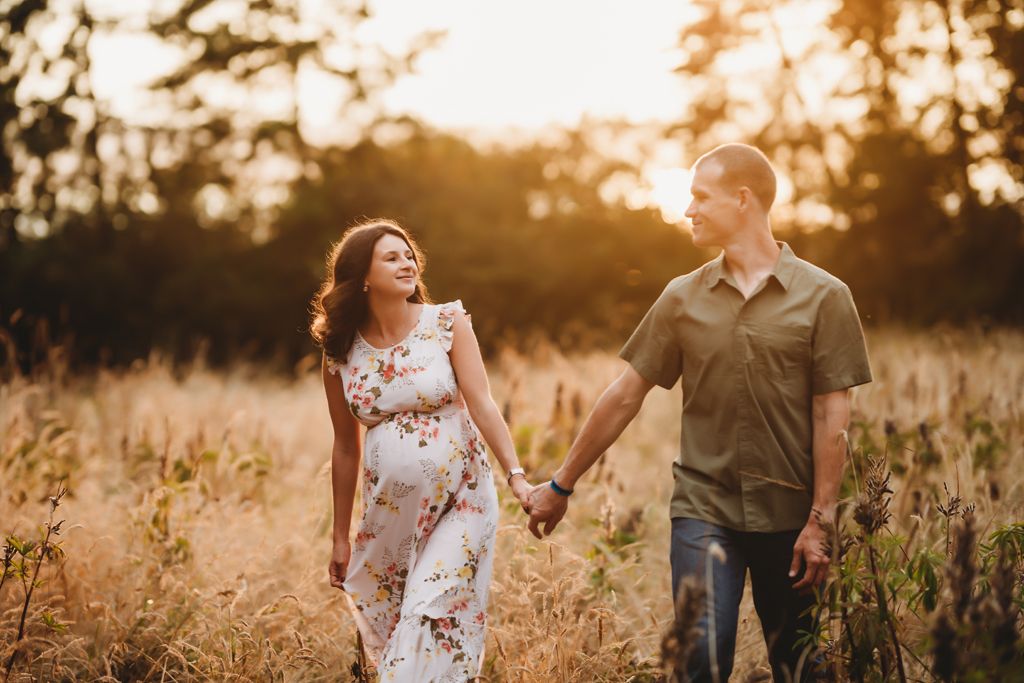 A couple walks hand in hand through a sunlit meadow during their maternity session.
