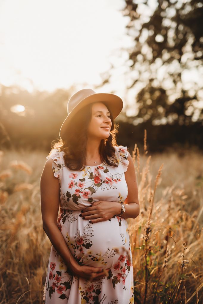 A pregnant woman in a hat and floral dress poses in a sunlit field.