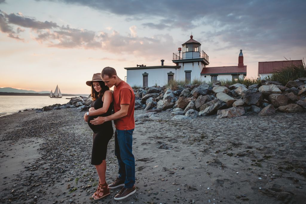 Couple standing close together near lighthouse at dusk with rocks and water in background.