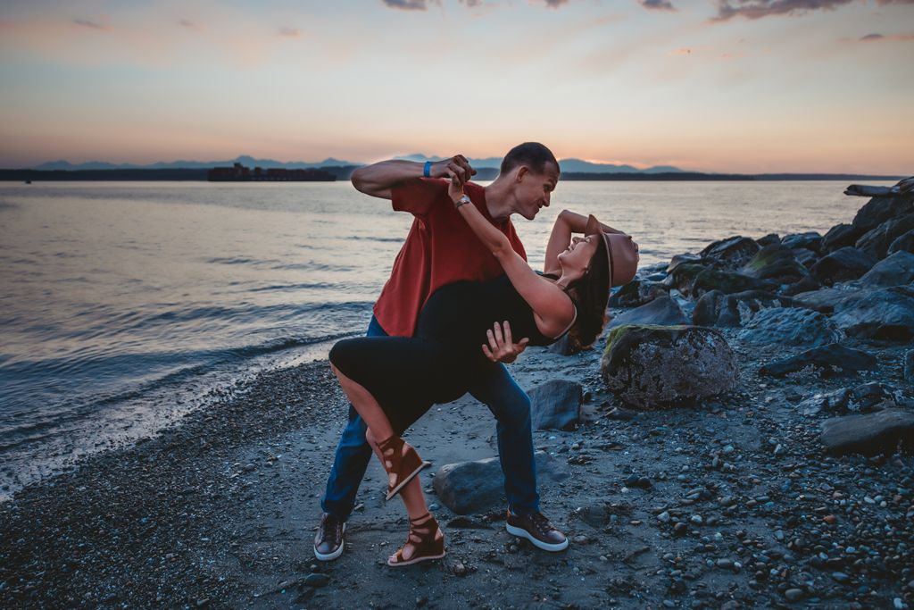 A romantic couple dances together on a rocky beach at sunset with mountains in the background.