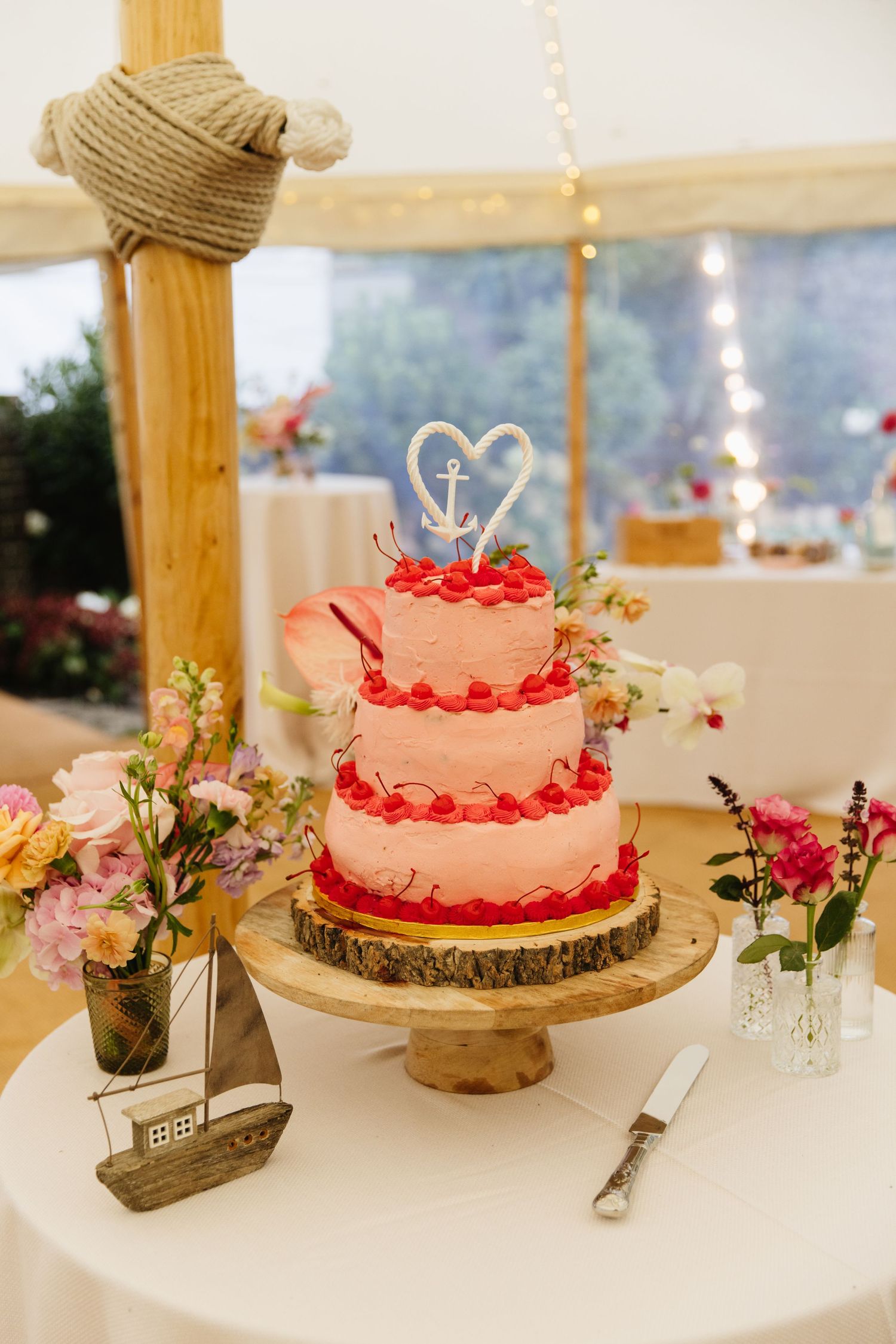 Three-tier white wedding cake with red accents and nautical details on rustic wood stand.