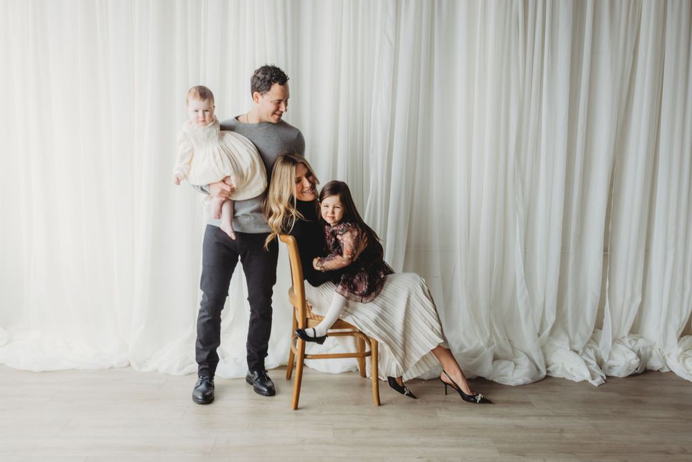 An elegant family portrait against a white curtain backdrop with a wooden chair and natural lighting.