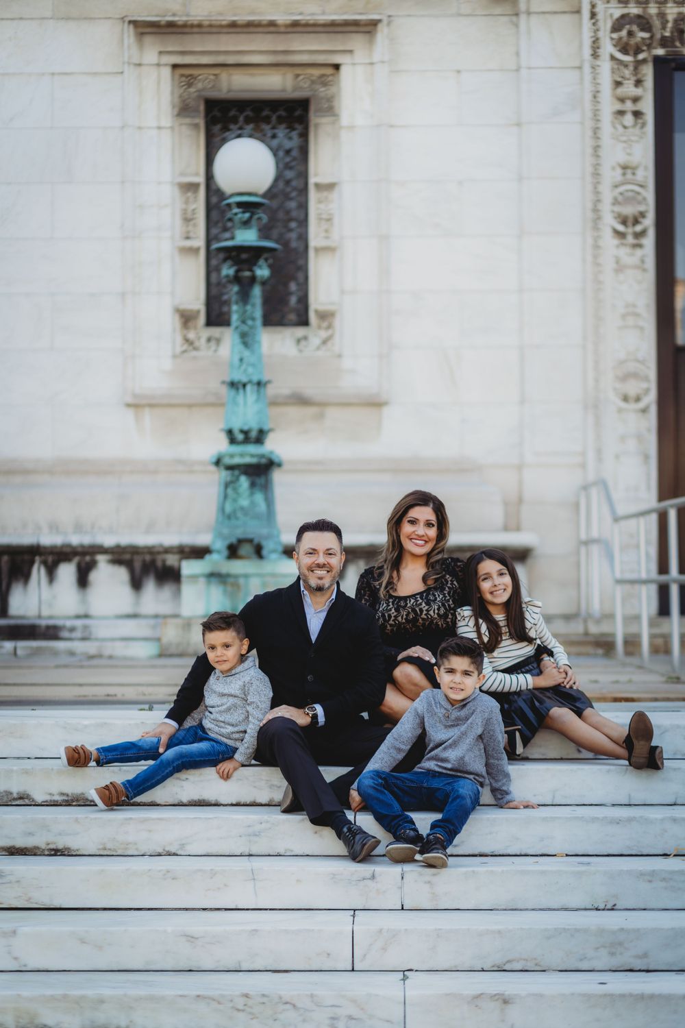 A family sits together on white stone steps near a decorative lamp post outside a building.