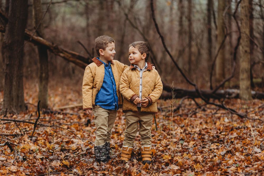 Two young children in matching tan coats stand together smiling in an autumn forest filled with fallen leaves.