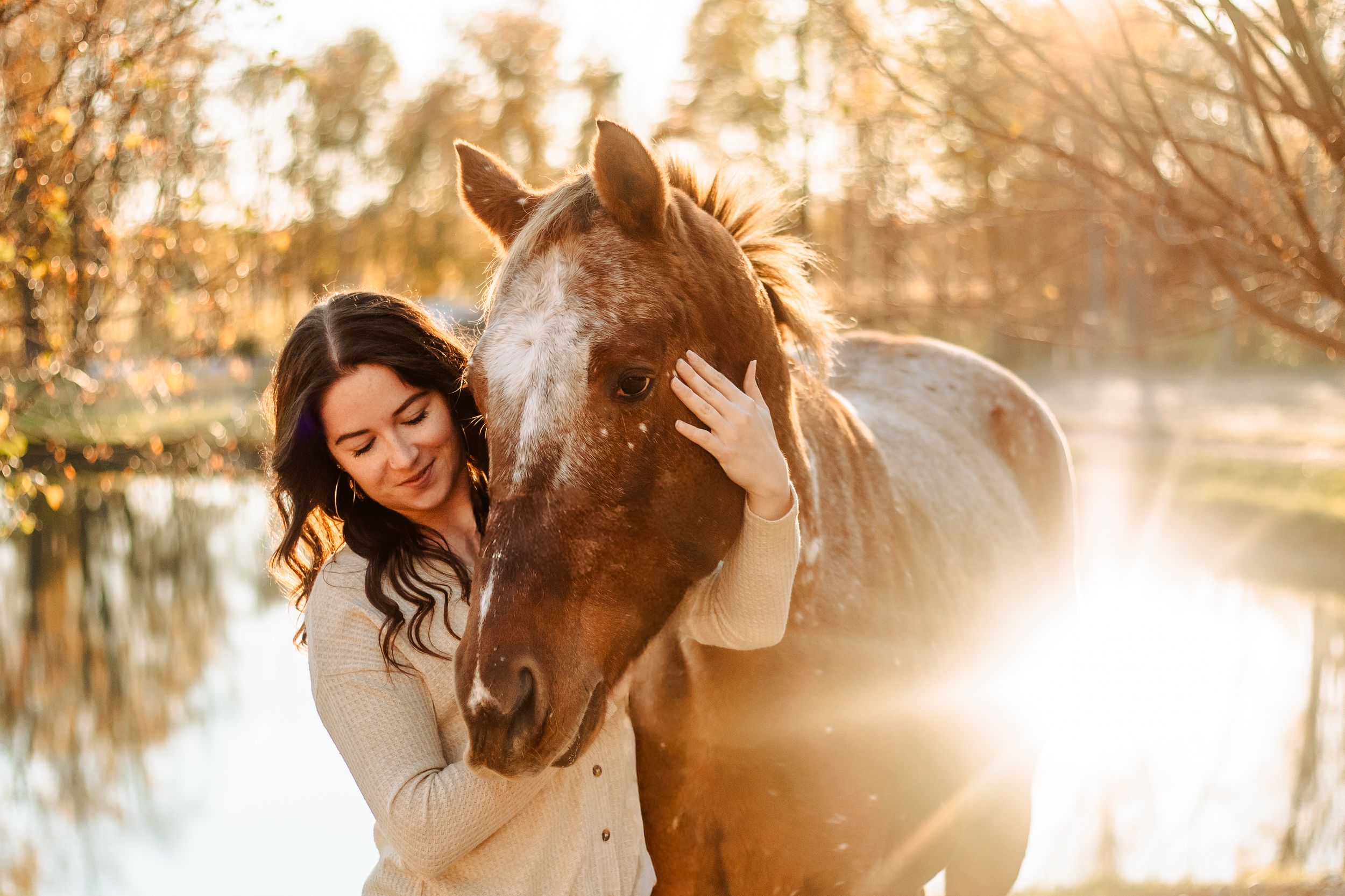 Woman in white dress poses with brown horse in sunlit forest setting during golden hour.