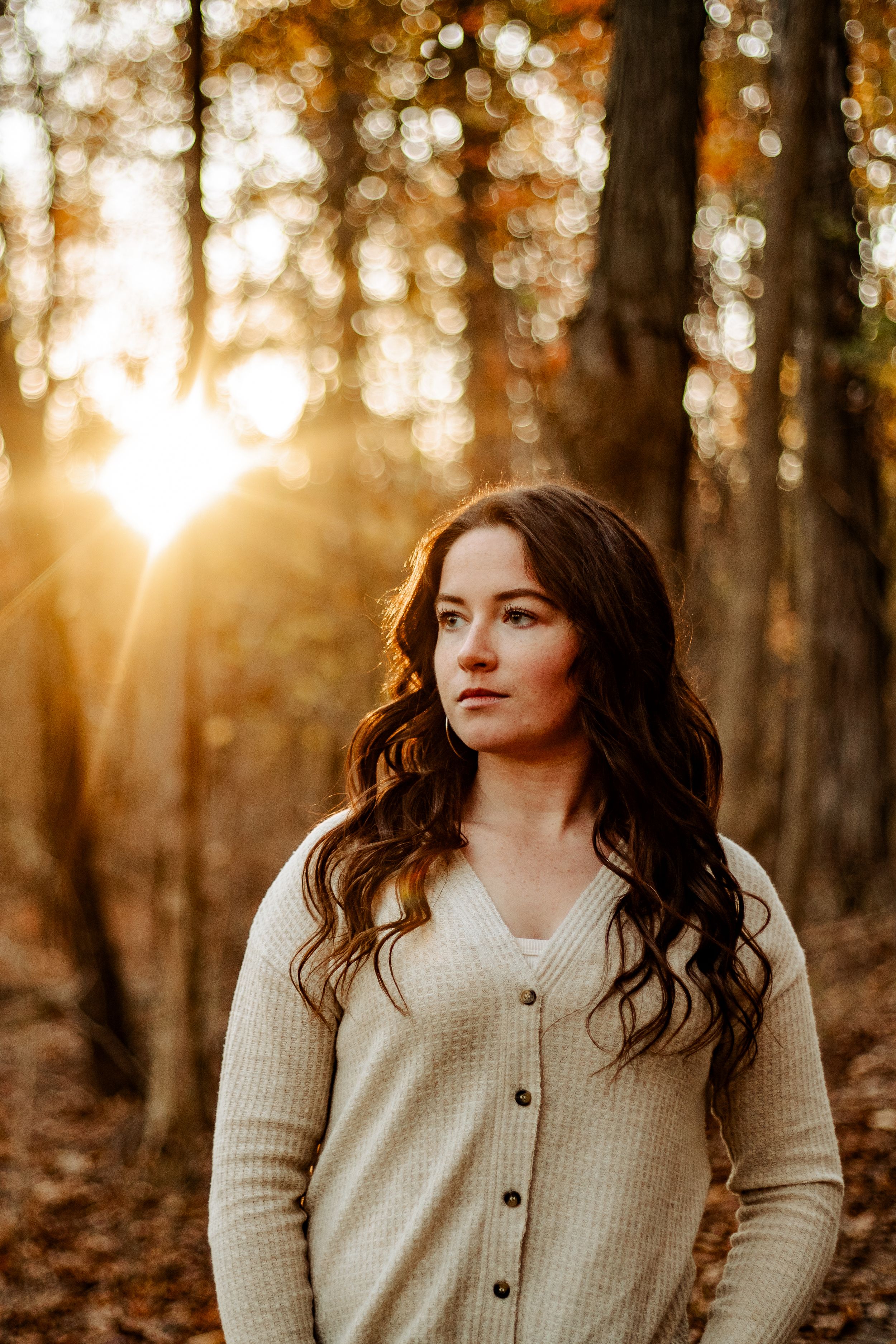 Portrait in autumn forest with golden sunlight filtering through trees as person in cream cardigan gazes into distance.