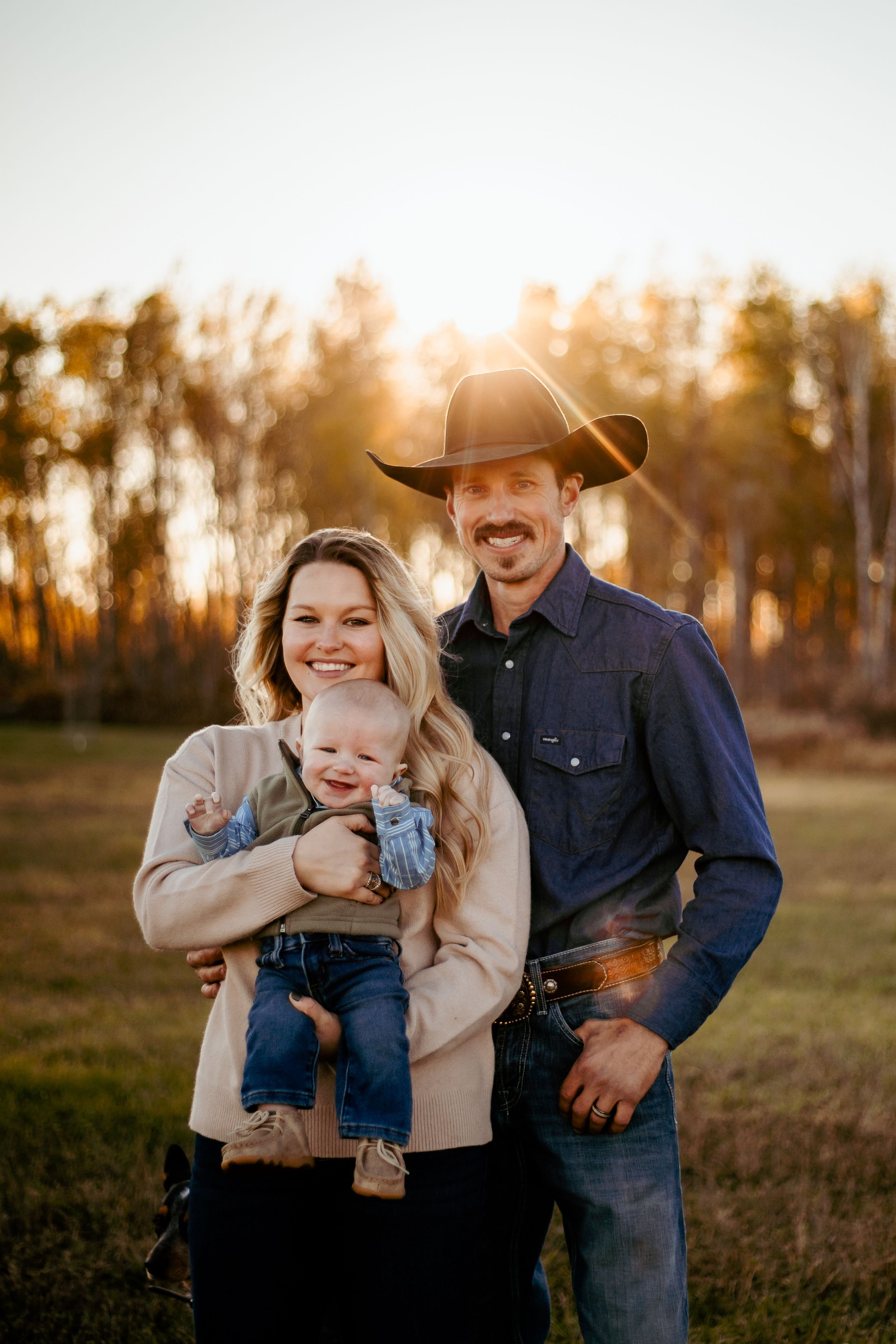 A couple and baby in western attire share joyful moments during a sunset photoshoot in a rustic field.