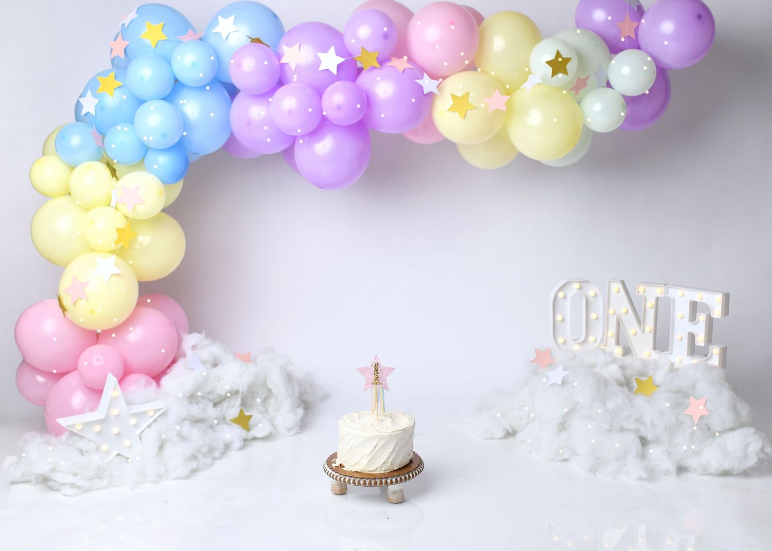 Pastel rainbow first birthday cake smash setup with purple, yellow, and blue balloons.