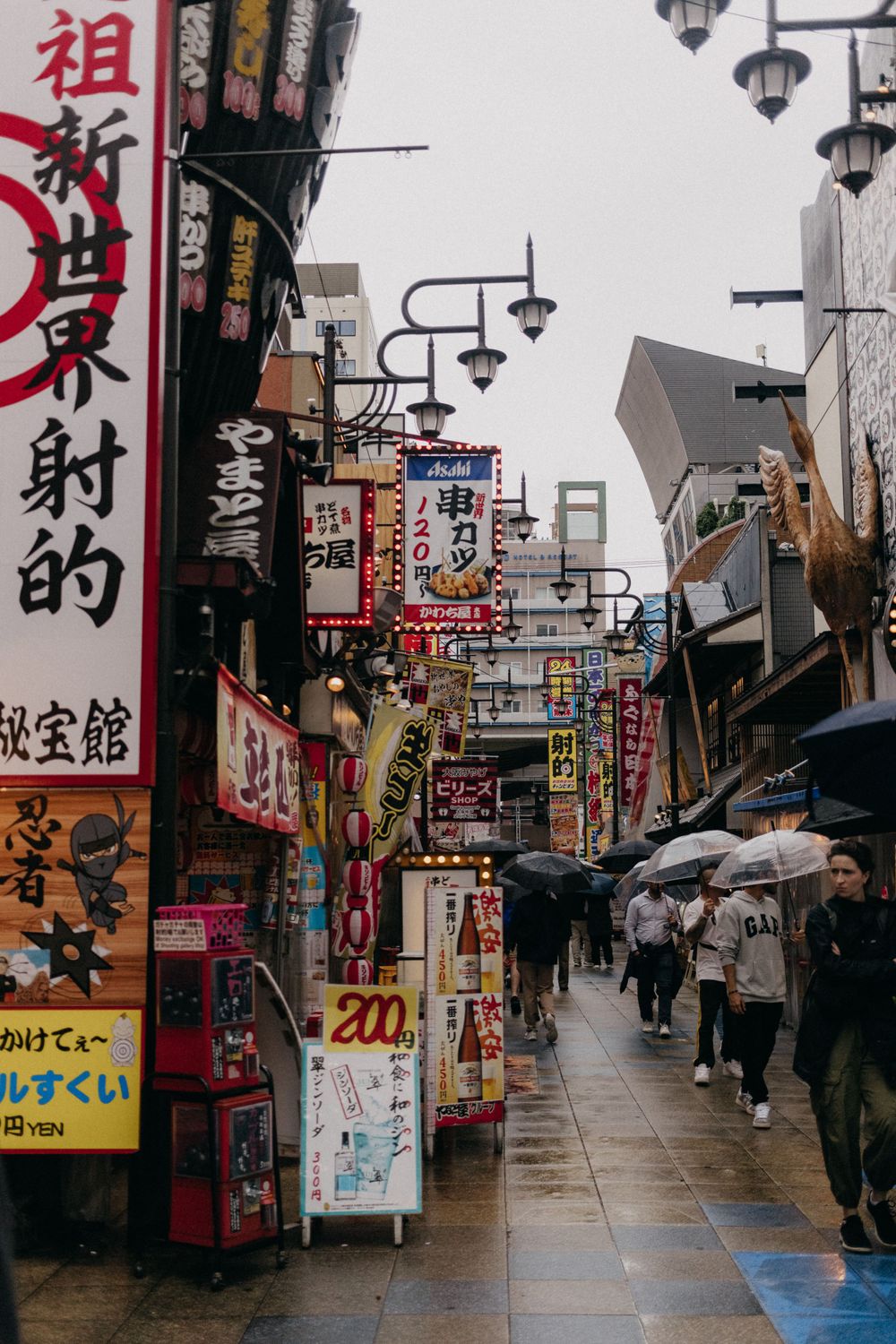 Busy Japanese street lined with colorful signs and umbrellas on a rainy day.